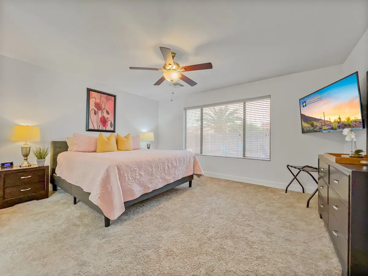 A spacious bedroom featuring a large bed with soft pink bedding, framed artwork above the headboard, a window with blinds allowing natural light to filter in, and a wall-mounted television. Nightstands with lamps frame the bed, contributing to the room's cozy and serene ambiance.