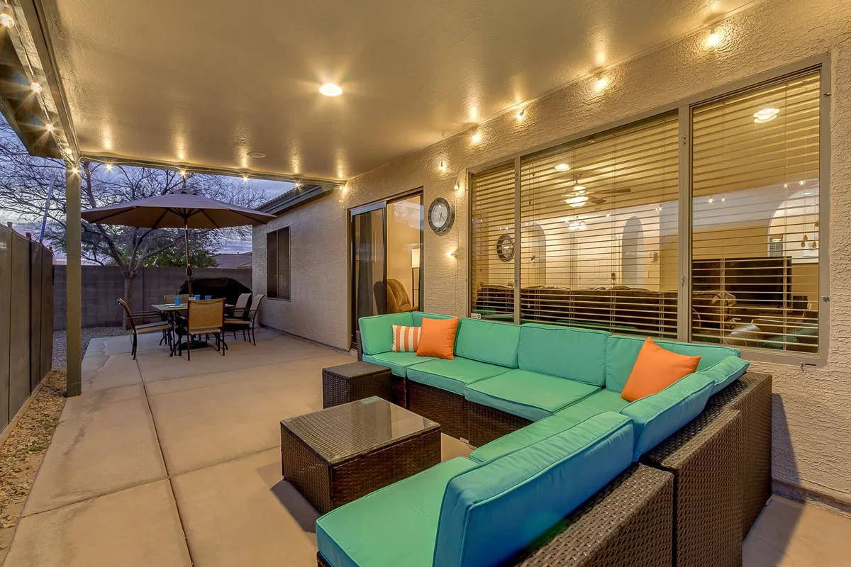 An exterior scene featuring a cozy turquoise sectional sofa, a large outdoor dining table under umbrellas, and glowing string lights. The space is designed for socializing, outdoor dining, and relaxing evenings under the stars.