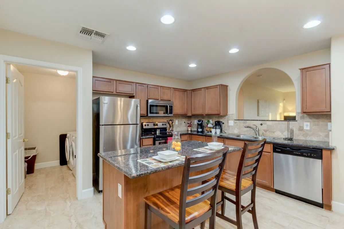 A bright, open-plan kitchen features a large island with bar stools, stainless steel appliances, warm wooden cabinetry, and a nearby dining table set for a meal. The layout flows toward a spacious lounge area, hinting at the seamless connection between cooking, dining, and relaxing spaces.