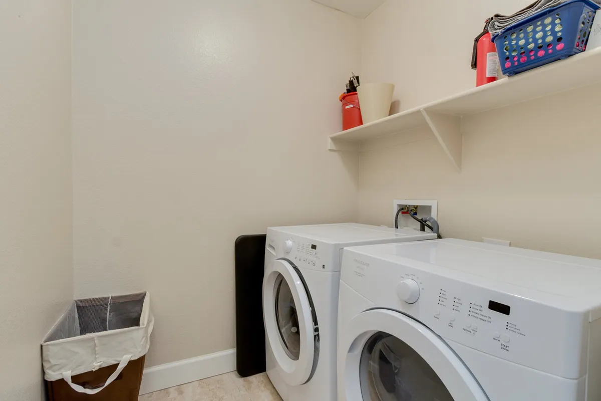 This image displays a functional laundry room, featuring a washing machine, dryer, shelf with various household items, and a waste bin. The setting is clean, organized, and embodies a sense of practical convenience for everyday living.