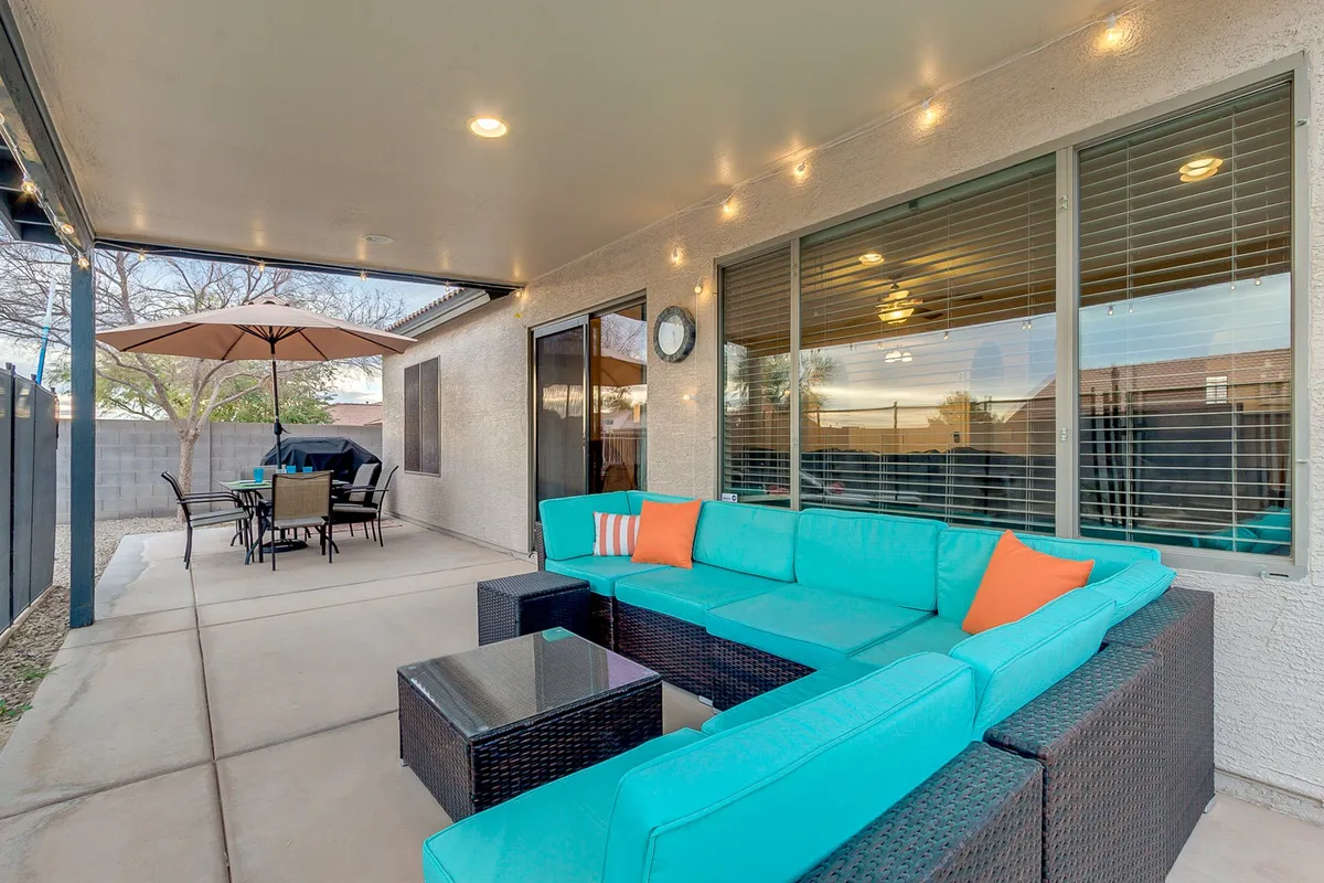 A stylish outdoor living area with a turquoise sectional sofa, a glass-top coffee table, and a dining set shaded by an umbrella. Large windows reflect the outdoor scene, and the atmosphere is casual and welcoming—ideal for group conversations or enjoying morning coffee together.
