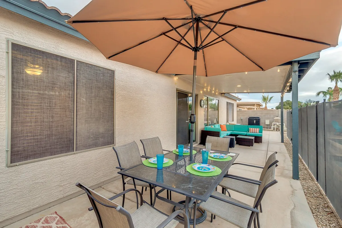 A cheerful outdoor dining area featuring a large table with an umbrella, surrounded by decorative pillows and ample seating. The setup is festive and inviting, perfect for hosting barbecue dinners or celebratory gatherings in a relaxed environment.