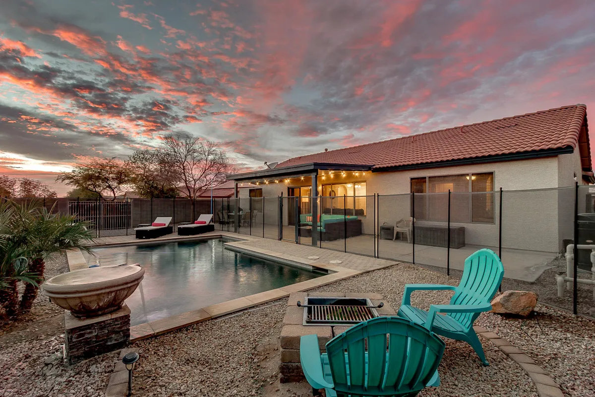 A beautiful backyard scene featuring a shimmering swimming pool surrounded by lounge chairs and a cozy outdoor seating area. The sky is lit with sunset hues, creating a tranquil, inviting atmosphere perfect for relaxing, swimming, or entertaining. The pool is securely fenced, ensuring privacy and peace of mind.
