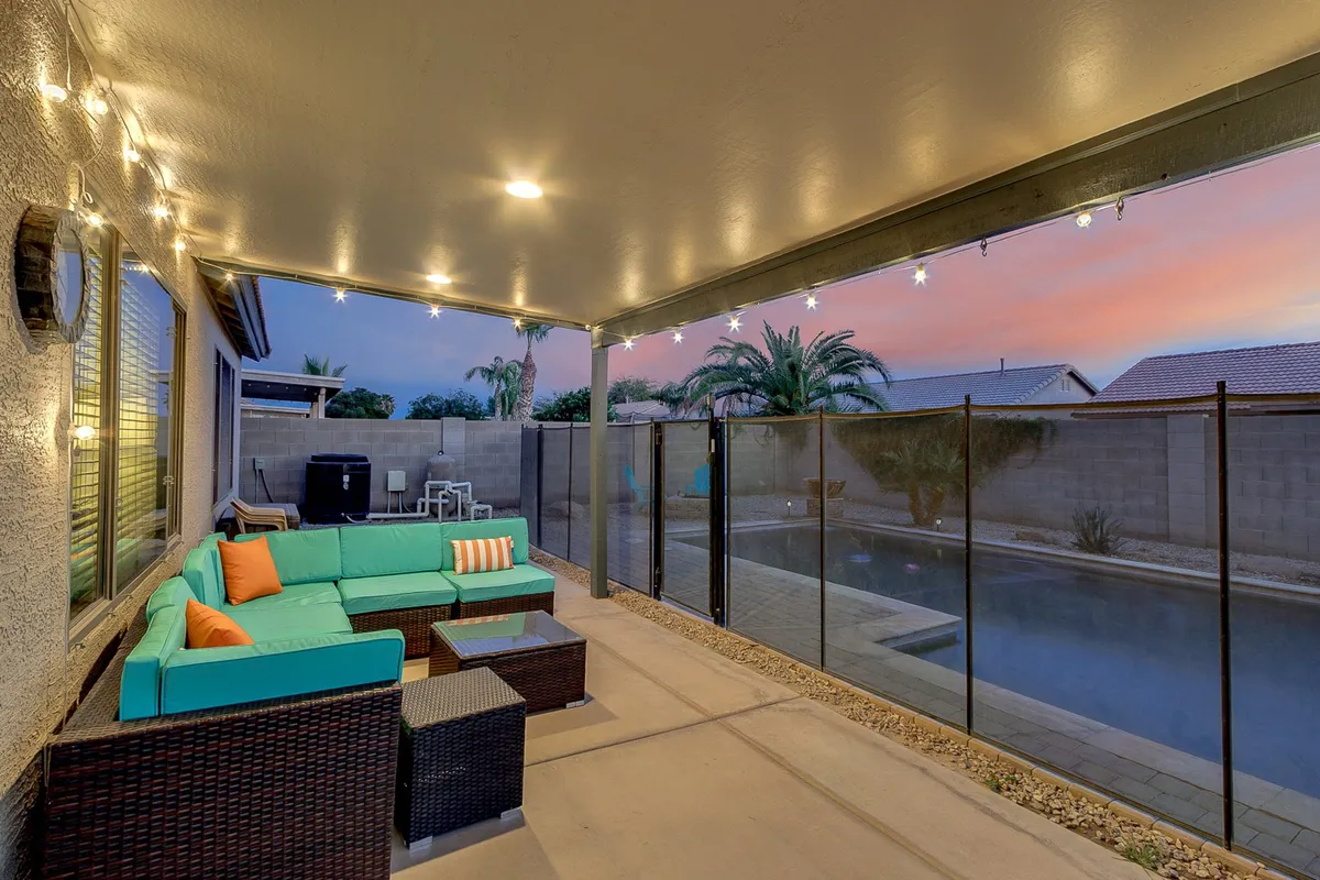 This exterior image showcases a tranquil backyard with a turquoise seating area, a sparkling swimming pool, string lights overhead, and lush palm trees. The setting exudes an inviting atmosphere ideal for evening gatherings and sunset relaxation.