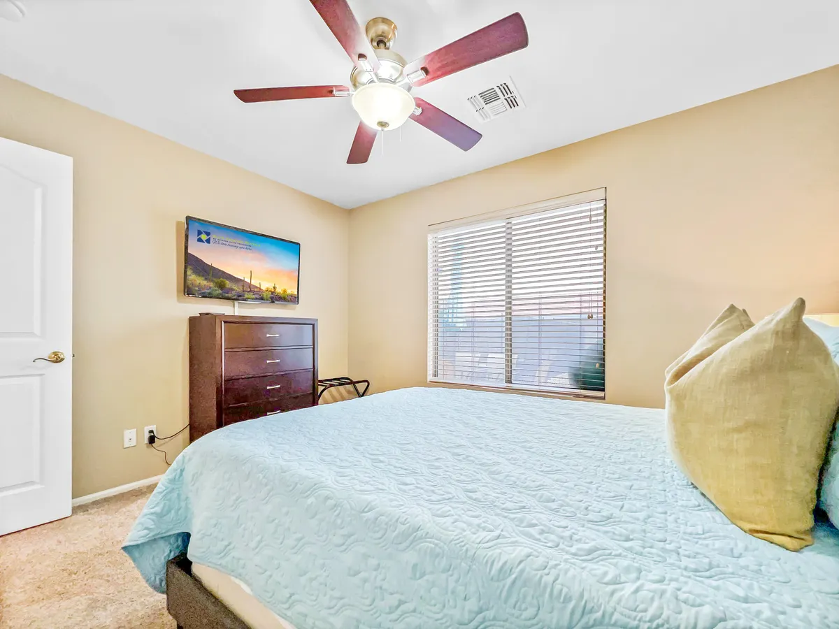 A cozy queen bedroom with a bed dressed in a light blue quilt, a brown dresser, a TV displaying a scenic landscape, and a window with blinds letting in gentle sunlight. The space feels welcoming and ideal for relaxation or enjoying a quiet moment in the morning or evening.