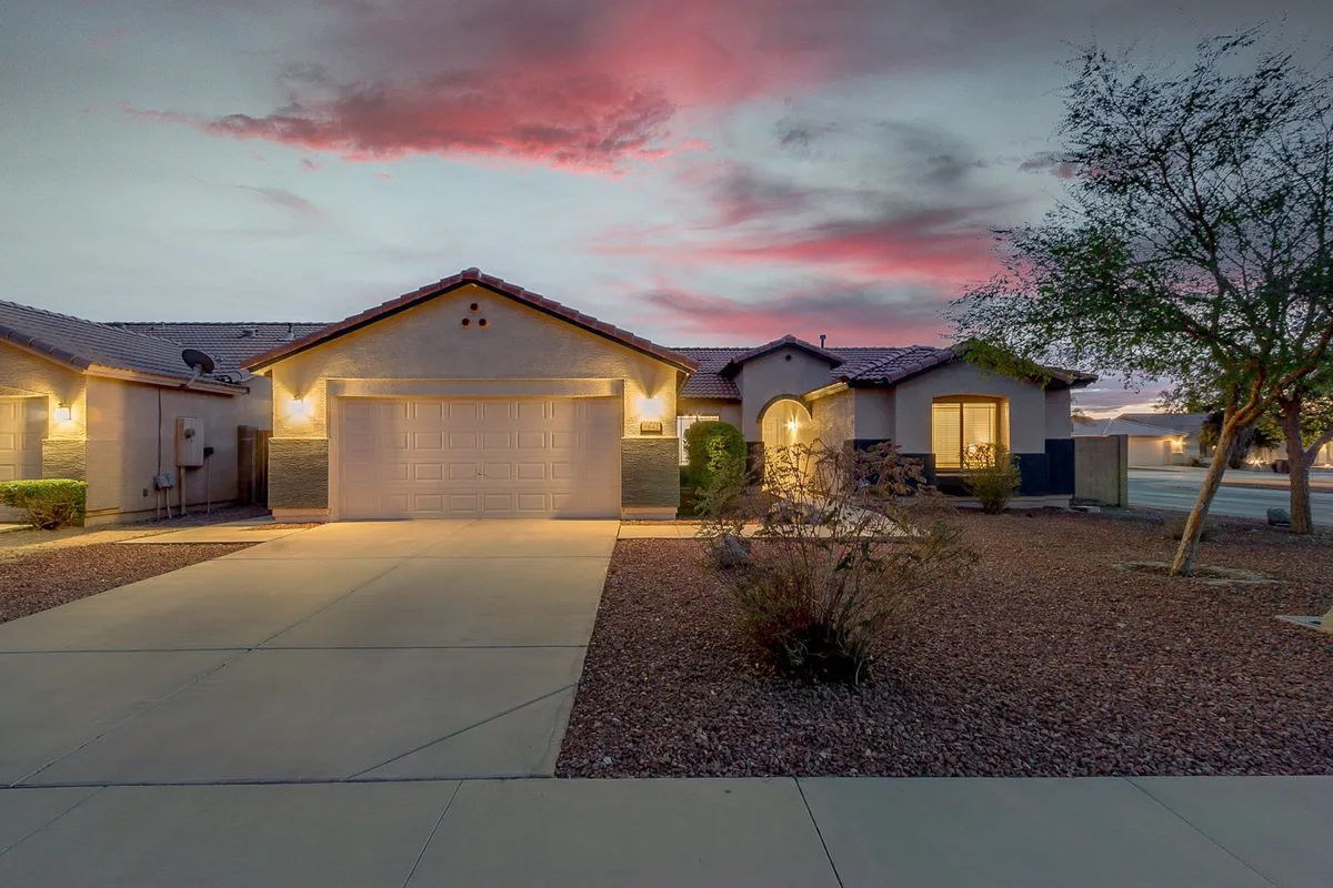 This image captures the front facade of the house with a landscaped garden and driveway. The inviting exterior is bathed in warm light, suggesting a welcoming atmosphere for residents returning from local markets or errands. The porch and pathway allude to quiet mornings or evenings spent outdoors, and the driveway connects directly to the mention of garage and street parking in the section.