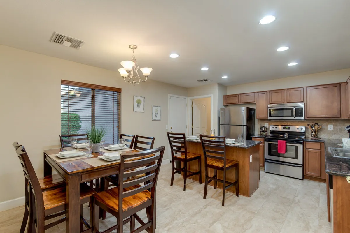 A large dining table sits adjacent to a kitchen island, with a modern chandelier overhead and a generous window letting in natural light. The area is designed for social dining, entertaining, and easy movement between kitchen and dining zones, reinforcing the sense of openness.